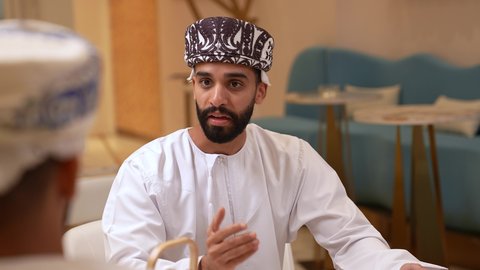 حوار بين اصدقاء في مطعم، تصوير مقرب لرجل عربي خليجي عماني يرتدي الدشداشة و الكمه يجلس يرفقه صديقة في المقهى, مناقشة مهام العمل في المطعم، التعارف وبناء علاقات اجتماعيه, صداقات ومعارف، الدفع والشراء من خلال برامج وتطبيقات الجوال