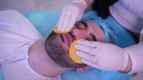 ترطيب وتقشير وتفتيح الجلد، استخدام مكونات تجميلية ، مفهوم العناية والإهتمام بنضارة وتحسين البشرة، طبيبة عربية خليجية عمانية تقوم بتنظيف الجلد باستخدام قطع اسفنجيه ، رجل عربي خليجي عماني يجلس على كرسي العيادة