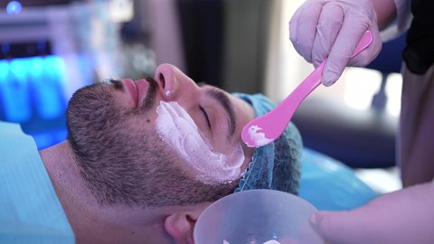 طبيبة مختصه فى العناية بالبشرة تضع الماسك على وجه الزبون ، مفهوم العناية والإهتمام بنضارة وتحسين البشرة، استخدام مكونات تجميلية شبابية، ترطيب وتنظيف وتفتيح الجلد، رجل عربي خليجي عماني يرتدي الدشداشة يجلس على كرسي العيادة