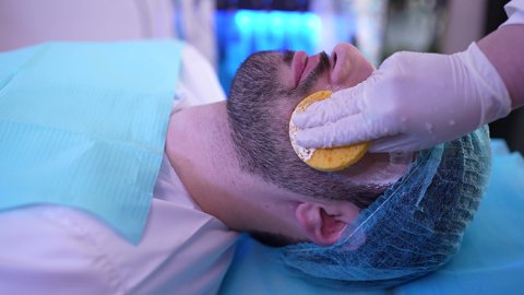 Using cosmetic ingredients to moisturize, exfoliate, and lighten the skin. The concept of skin care and attention to freshness and improvement. An Arab Gulf Omani female doctor cleansing the skin using sponges. An Arab Gulf Omani male sitting in a clinic chair.