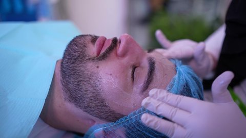 مفهوم العناية والإهتمام بنضارة وتحسين البشرة، استخدام مكونات تجميلية شبابية، ترطيب وتنظيف وتفتيح الجلد، رجل عربي خليجي عماني يرتدي الدشداشة يجلس على كرسي العيادة ، طبيبة مختصه فى العناية بالبشرة تضع كريم المرطب على وجه الزبون