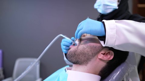 شاب عربي خليجي عماني يرتدي الدشداشة والكمة يجلس على كرسي العلاج في العيادة السنية ، الزيارة الدورية للاطمئنان على الصحة الفموية، معالجة النخور وتركيب الحشوات، استخدام المعدات اللازمة لجراحة الفم، تصوير مقرب من الجانب لطبيبة ترتدي القفازات تقوم بفحص أسنان المريض وتشخيص المرض