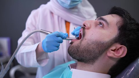 تصوير مقرب من الجانب لطبيبة ترتدي القفازات تقوم بفحص أسنان المريض وتشخيص المرض، شاب عربي خليجي عماني يرتدي الدشداشة والكمة يجلس على كرسي العلاج في العيادة السنية ، الزيارة الدورية للاطمئنان على الصحة الفموية، معالجة النخور وتركيب الحشوات، استخدام المعدات اللازمة لجراحة الفم