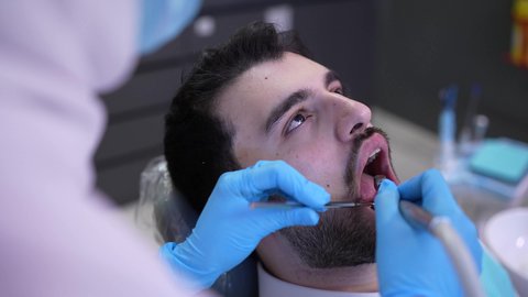 معالجة النخور وتركيب الحشوات،  استخدام المعدات اللازمة لجراحة الفم, الزيارة الدورية للاطمئنان على الصحة الفموية، تصوير مقرب من الجانب لطبيبة ترتدي القفازات تقوم بفحص أسنان المريض وتشخيص المرض، شاب عربي خليجي عماني يرتدي الدشداشة والكمة يجلس على كرسي العلاج في العيادة السنية