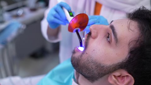 مفهوم الطب والرعاية الصحية، استخدام المعدات اللازمة لجراحة الاسنان ، تصوير مقرب من الجانب لطبيب يرتدي القفازات يقوم بفحص أسنان المريض، شاب عربي خليجي عماني يرتدي الدشداشة والكمة يجلس على كرسي العلاج في العيادة السنية، تقديم الخدمات الطبية للمرضى ، إجراء الفحوصات الدورية للعناية بالصحة الفموية