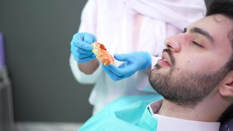 تقديم نصائح للمحافظة على صحة الفم والأسنان، مفهوم الطب والرعاية الصحية، طبيبة أسنان ترتدي المعطف والقفازات الطبية تمسك بيدها نموذج سني، الاهتمام في الصحة الفموية ،  تصوير مقرب لرجل عربي خليجي عماني يرتدي الكمه يجلس على كرسي العلاج