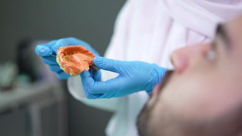 طبيبة أسنان ترتدي المعطف والقفازات الطبية تمسك بيدها نموذج سني، الاهتمام في الصحة الفموية ،  تصوير مقرب لرجل عربي خليجي عماني يرتدي الكمه يجلس على كرسي العلاج ، تقديم نصائح للمحافظة على صحة الفم والأسنان، مفهوم الطب والرعاية الصحية