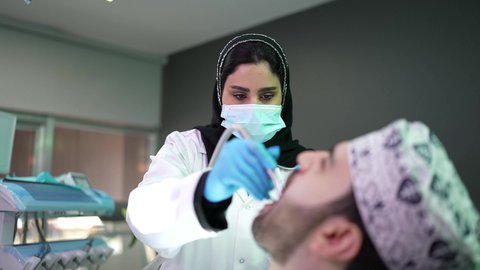 استخدام المعدات اللازمة لجراحة الفم ، معالجة النخور وتركيب الحشوات ، شاب عربي خليجي عماني يرتدي الدشداشة والكمة يجلس على كرسي العلاج في العيادة السنية، تصوير مقرب من الجانب لطبيبة ترتدي القفازات تقوم بفحص أسنان المريض وتشخيص المرض، الزيارة الدورية للاطمئنان على الصحة الفموية