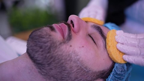 استخدام مكونات تجميلية ، ترطيب وتقشير وتفتيح الجلد، مفهوم العناية والإهتمام بنضارة وتحسين البشرة، طبيبة عربية خليجية عمانية تقوم بتنظيف الجلد باستخدام قطع اسفنجيه ، رجل عربي خليجي عماني يجلس على كرسي العيادة