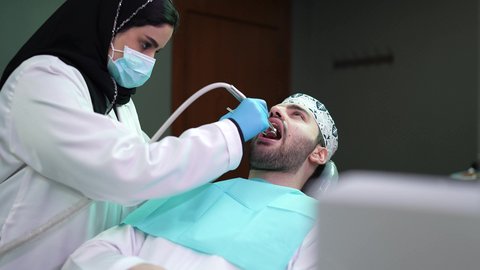 معالجة النخور وتركيب الحشوات ، شاب عربي خليجي عماني يرتدي الدشداشة والكمة يجلس على كرسي العلاج في العيادة السنية، تصوير مقرب من الجانب لطبيبة ترتدي القفازات تقوم بفحص أسنان المريض وتشخيص المرض، الزيارة الدورية للاطمئنان على الصحة الفموية، استخدام المعدات اللازمة لجراحة الفم
