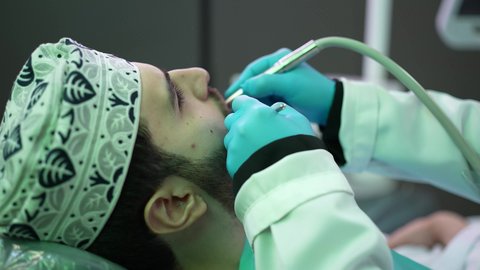 الزيارة الدورية للاطمئنان على الصحة الفموية، استخدام المعدات اللازمة لجراحة الفم, معالجة النخور وتركيب الحشوات، تصوير مقرب من الجانب لطبيبة ترتدي القفازات تقوم بفحص أسنان المريض وتشخيص المرض، شاب عربي خليجي عماني يرتدي الدشداشة والكمة يجلس على كرسي العلاج في العيادة السنية