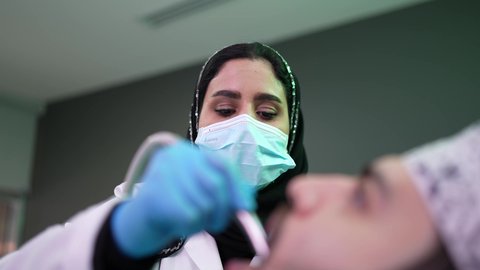 معالجة النخور وتركيب الحشوات، استخدام المعدات اللازمة لجراحة الفم, الزيارة الدورية للاطمئنان على الصحة الفموية، تصوير مقرب من الجانب لطبيبة ترتدي القفازات تقوم بفحص أسنان المريض وتشخيص المرض، شاب عربي خليجي عماني يرتدي الدشداشة والكمة يجلس على كرسي العلاج في العيادة السنية