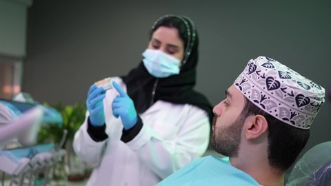 طبيبة أسنان ترتدي المعطف والقفازات الطبية تمسك بيدها نموذج سني، الاهتمام في الصحة الفموية ،  تصوير مقرب لرجل عربي خليجي عماني يرتدي الكمه يجلس على كرسي العلاج ، تقديم نصائح للمحافظة على صحة الفم والأسنان، مفهوم الطب والرعاية الصحية
