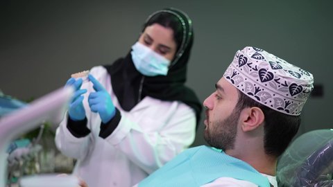 تصوير مقرب لرجل عربي خليجي عماني يرتدي الكمه يجلس على كرسي العلاج ، طبيبة أسنان ترتدي المعطف والقفازات الطبية تمسك بيدها نموذج سني، الاهتمام في الصحة الفموية ،  تقديم نصائح للمحافظة على صحة الفم والأسنان، مفهوم الطب والرعاية الصحية