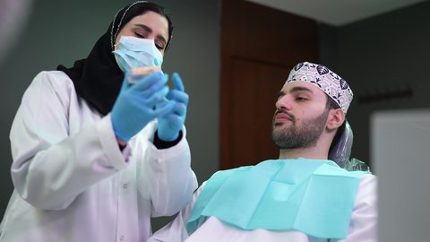 تقديم نصائح للمحافظة على صحة الفم والأسنان، مفهوم الطب والرعاية الصحية, الاهتمام في الصحة الفموية ، تصوير مقرب لرجل عربي خليجي عماني يرتدي الكمه يجلس على كرسي العلاج ، طبيبة أسنان ترتدي المعطف والقفازات الطبية تمسك بيدها نموذج سني