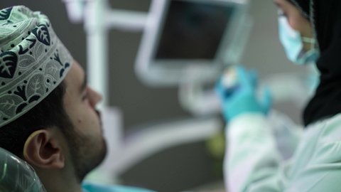 الاهتمام في الصحة الفموية ، تصوير مقرب لرجل عربي خليجي عماني يرتدي الكمه يجلس على كرسي العلاج ، طبيبة أسنان ترتدي المعطف والقفازات الطبية تمسك بيدها نموذج سني،مفهوم الطب والرعاية الصحية, تقديم نصائح للمحافظة على صحة الفم والأسنان