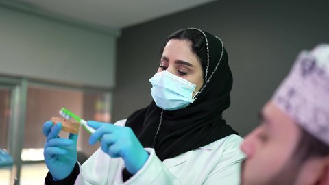 طبيبة أسنان ترتدي المعطف والقفازات الطبية تمسك بيدها نموذج سني،مفهوم الطب والرعاية الصحية, تقديم نصائح للمحافظة على صحة الفم والأسنان، الاهتمام في الصحة الفموية,  تصوير مقرب لرجل عربي خليجي عماني يرتدي الكمه يجلس على كرسي العلاج