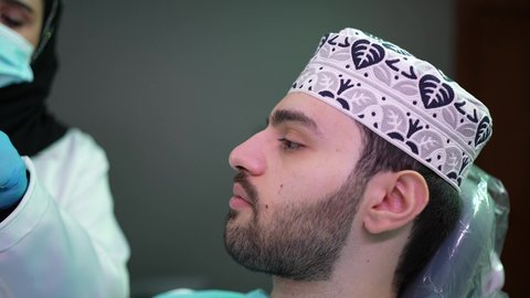 مفهوم الطب والرعاية الصحية, تقديم نصائح للمحافظة على صحة الفم والأسنان، الاهتمام في الصحة الفموية, طبيبة أسنان ترتدي المعطف والقفازات الطبية تمسك بيدها نموذج سني، تصوير مقرب لرجل عربي خليجي عماني يرتدي الكمه يجلس على كرسي العلاج