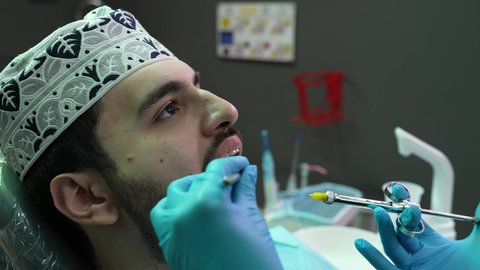 تقديم الرعاية الصحية في المراكز التخصصية،  الاستعداد لحقن البنج الموضعي لتخفيف الشعور بالألم ,تصوير مقرب لرجل عربي خليجي عماني يرتدي الزي التقليدي يجلس في العيادة السنية بإيماءات السرور, يد طبيب أسنان يرتدي القفازات الطبية يمسك بيده إبرة المخدر،معالجة النخور والتسوس