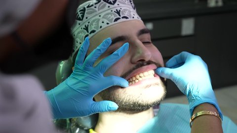 مفهوم الطب والرعاية الصحية، تصوير مقرب من الجانب لطبيب يرتدي القفازات يقوم بفحص أسنان المريض، شاب عربي خليجي عماني يرتدي الدشداشة والكمة يجلس على كرسي العلاج في العيادة السنية، تقديم الخدمات الطبية للمرضى ، إجراء الفحوصات الدورية للعناية بالصحة الفموية