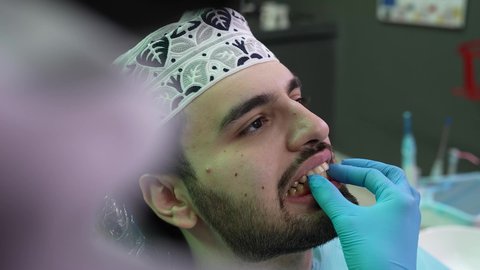 تصوير مقرب من الجانب لطبيب يرتدي القفازات يقوم بفحص أسنان المريض، شاب عربي خليجي عماني يرتدي الدشداشة والكمة يجلس على كرسي العلاج في العيادة السنية، تقديم الخدمات الطبية للمرضى ، إجراء الفحوصات الدورية للعناية بالصحة الفموية، مفهوم الطب والرعاية الصحية