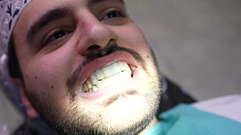 تقديم الخدمات الطبية للمرضى ، إجراء الفحوصات الدورية للعناية بالصحة الفموية، مفهوم الطب والرعاية الصحية، تصوير مقرب من الجانب لطبيب يرتدي القفازات يقوم بفحص أسنان المريض، شاب عربي خليجي عماني يرتدي الدشداشة والكمة يجلس على كرسي العلاج في العيادة السنية