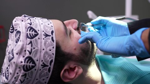 تصوير مقرب من الجانب لطبيب يرتدي القفازات يقوم بفحص أسنان المريض، شاب عربي خليجي عماني يرتدي الدشداشة والكمة يجلس على كرسي العلاج في العيادة السنية، إجراء الفحوصات الدورية للعناية بالصحة الفموية، تقديم الخدمات الطبية للمرضى,مفهوم الطب والرعاية الصحية