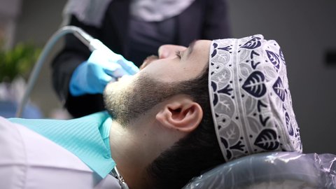 تقديم الخدمات الطبية للمرضى,مفهوم الطب والرعاية الصحية، إجراء الفحوصات الدورية للعناية بالصحة الفموية، تصوير مقرب من الجانب لطبيب يرتدي القفازات يقوم بفحص أسنان المريض، شاب عربي خليجي عماني يرتدي الدشداشة والكمة يجلس على كرسي العلاج في العيادة السنية