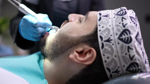 مفهوم الطب والرعاية الصحية، تقديم الخدمات الطبية للمرضى, إجراء الفحوصات الدورية للعناية بالصحة الفموية، تصوير مقرب من الجانب لطبيب يرتدي القفازات يقوم بفحص أسنان المريض، شاب عربي خليجي عماني يرتدي الدشداشة والكمة يجلس على كرسي العلاج في العيادة السنية
