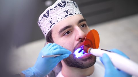 إجراء الفحوصات الدورية للعناية بالصحة الفموية ، استخدام المعدات اللازمة لجراحة الاسنان ، مفهوم الطب والرعاية الصحية، تصوير مقرب من الجانب لطبيب يرتدي القفازات يقوم بفحص أسنان المريض، شاب عربي خليجي عماني يرتدي الدشداشة والكمة يجلس على كرسي العلاج في العيادة السنية، تقديم الخدمات الطبية للمرضى