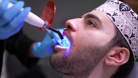 استخدام المعدات اللازمة لجراحة الاسنان ، مفهوم الطب والرعاية الصحية، تصوير مقرب من الجانب لطبيب يرتدي القفازات يقوم بفحص أسنان المريض، شاب عربي خليجي عماني يرتدي الدشداشة والكمة يجلس على كرسي العلاج في العيادة السنية، تقديم الخدمات الطبية للمرضى ، إجراء الفحوصات الدورية للعناية بالصحة الفموية