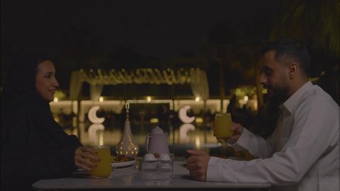 Spending a pleasant time outdoors in a pleasant atmosphere, drinking fresh juices, a Saudi Arabian Gulf couple sitting in a café, exchanging conversations and discussions, the concept of a happy family, a weekend getaway, going out for a family evening.