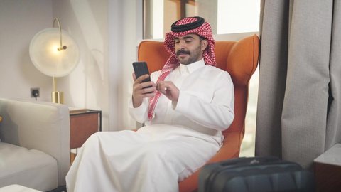 إجراء اتصال هاتفي عبر الجوال، الدردشه عبر الانترنت ،مهن و أعمال في المملكه العربيه السعوديه، شاب عربي خليجي سعودي يرتدي الزى التقليدي والشماغ يمسك الهاتف المحمول بيده ، يجلس على  كرسي مريح فى غرفه فندقيه ، تصفح وسائل التواصل الاجتماعي ،  دمج التقنيات المتطورة في الحياه اليوميه
