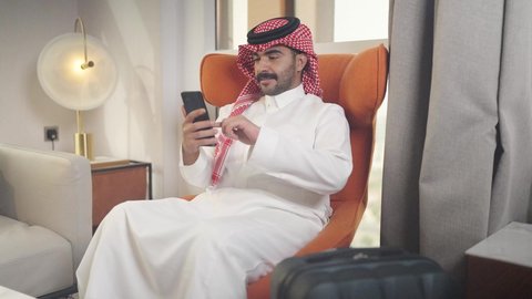 شاب عربي خليجي سعودي يرتدي الزى التقليدي والشماغ يمسك الهاتف المحمول بيده ، يجلس على  كرسي مريح فى غرفه فندقيه ، تصفح وسائل التواصل الاجتماعي ،  دمج التقنيات المتطورة في الحياه اليوميه ،إجراء اتصال هاتفي عبر الجوال، الدردشه عبر الانترنت ،مهن و أعمال في المملكه العربيه السعوديه