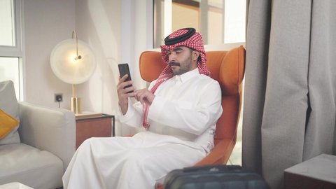 تصفح وسائل التواصل الاجتماعي ،  دمج التقنيات المتطورة في الحياه اليوميه ،إجراء اتصال هاتفي عبر الجوال، الدردشه عبر الانترنت ، شاب عربي خليجي سعودي يرتدي الزى التقليدي والشماغ يمسك الهاتف المحمول بيده ، يجلس على  كرسي مريح فى غرفه فندقيه ، مهن و أعمال في المملكه العربيه السعوديه