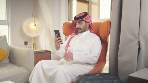 الدردشه عبر الانترنت ،  دمج التقنيات المتطورة في الحياه اليوميه ،إجراء اتصال هاتفي عبر الجوال، تصفح وسائل التواصل الاجتماعي ، شاب عربي خليجي سعودي يرتدي الزى التقليدي والشماغ يمسك الهاتف المحمول بيده ، يجلس على  كرسي مريح فى غرفه فندقيه ، مهن و أعمال في المملكه العربيه السعوديه