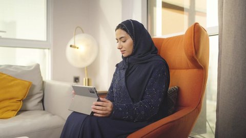 Using modern technology for work, the concept of remote project management, a veiled Saudi Arabian Gulf woman wearing a black abaya completing work tasks via a tablet, secure online shopping, an Omani woman sitting on a chair in a hotel room