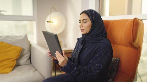 مفهوم إدارة المشاريع عن بعد, إمرأة عربية خليجية سعودية محجبة ترتدي عباءة سوداء تقوم بانجاز مهام العمل عبر جهاز التابلت, إستخدام اجهزة تقنية حديثة للعمل، التسوق الإلكتروني الآمن ، سيده عمانيه تجلس على كرسي فى غرفه فندقيه