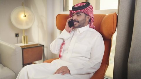 دمج التقنيات المتطورة في الحياه اليوميه ،إجراء اتصال هاتفي عبر الجوال، الدردشه عبر الانترنت ، تصفح وسائل التواصل الاجتماعي ، شاب عربي خليجي سعودي يرتدي الزى التقليدي والشماغ يمسك الهاتف المحمول بيده ، يجلس على  كرسي مريح فى غرفه فندقيه ، مهن و أعمال في المملكه العربيه السعوديه