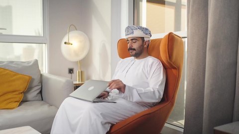 وظائف ومهن سعودية ، إنجاز المهام المطلوبة عبر اللابتوب، ادارة الأعمال عن بعد، دمج التقنيات المتطوره فى الحياه اليوميه ،التطور والتكنولوجيا الحديثة، رجل عربي خليجي سعودي يرتدي الزى التقليدي مع الشماغ يجلس على كرسي مريح في غرفة فندقيه ويستخدم جهاز الحاسوب المحمول