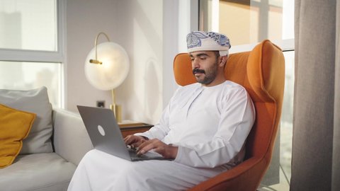 اجراء مكالمه فيديو، إنجاز المهام المطلوبة عبر اللابتوب، ادارة الأعمال عن بعد، دمج التقنيات المتطوره فى الحياه اليوميه ،التطور والتكنولوجيا الحديثة، رجل عربي خليجي سعودي يرتدي الزى التقليدي مع الشماغ يجلس على كرسي مريح في غرفة فندقيه ويستخدم جهاز الحاسوب المحمول ،وظائف ومهن سعودية