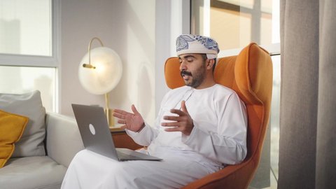 إنجاز المهام المطلوبة عبر اللابتوب، ادارة الأعمال عن بعد، دمج التقنيات المتطوره فى الحياه اليوميه ،التطور والتكنولوجيا الحديثة، رجل عربي خليجي سعودي يرتدي الزى التقليدي مع الشماغ يجلس على كرسي مريح في غرفة فندقيه ويستخدم جهاز الحاسوب المحمول ،وظائف ومهن سعودية