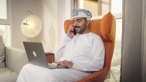 التطور والتكنولوجيا الحديثة،  ادارة الأعمال عن بعد، اجراء مكالمه هاتفيه، دمج التقنيات المتطوره فى الحياه اليوميه ، إنجاز المهام المطلوبة عبر اللابتوب، رجل عربي خليجي سعودي يرتدي الزى التقليدي مع الشماغ يجلس على كرسي مريح في غرفة فندقيه ويستخدم جهاز الحاسوب المحمول والجوال ،وظائف ومهن سعودية