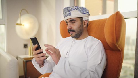 الدردشه عبر الانترنت ، دمج التقنيات المتطورة في الحياه اليوميه ، تصفح وسائل التواصل الاجتماعي ، شاب عربي خليجي عماني يرتدي الدشداشه والكمه يمسك الهاتف المحمول بيده ، يجلس على  كرسي مريح فى غرفه فندقيه ، إجراء اتصال هاتفي عبر الجوال،  اجراء مكالمه فيديو ، مهن و أعمال في سلطنه عمان