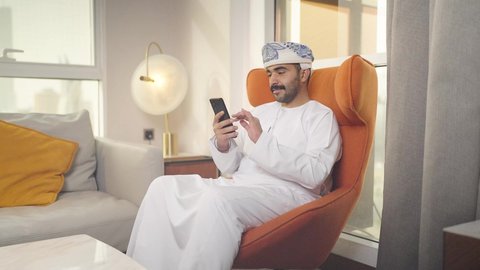 اجراء بث مباشر ،الدردشه عبر الانترنت ، دمج التقنيات المتطورة في الحياه اليوميه ، تصفح وسائل التواصل الاجتماعي ، شاب عربي خليجي عماني يرتدي الدشداشه والكمه يمسك الهاتف المحمول بيده ، يجلس على  كرسي مريح فى غرفه فندقيه ، إجراء اتصال هاتفي عبر الجوال، مهن و أعمال في سلطنه عمان