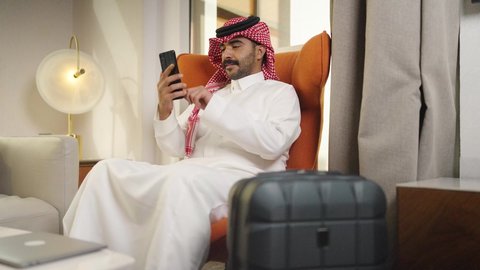 Making a phone call via mobile phone, using modern and advanced technologies at work, browsing social media, communicating with clients remotely, making a video call, professions and businesses in the Sultanate of Oman, a young Arab Gulf Omani man wearing a dishdasha and a cloak holding a mobile phone, sitting on a comfortable chair in a hotel room.