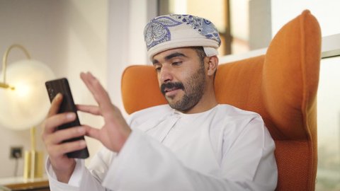 شاب عربي خليجي عماني يرتدي الدشداشه والكمه يمسك الهاتف المحمول بيده ، يجلس على  كرسي مريح فى غرفه فندقيه ، إجراء اتصال هاتفي عبر الجوال، استخدام التقنيات الحديثة و المتطورة في العمل ،  تصفح وسائل التواصل الاجتماعي ، التواصل مع العملاء عن بعد، اجراء مكالمه فيديو ، مهن و أعمال في سلطنه عمان