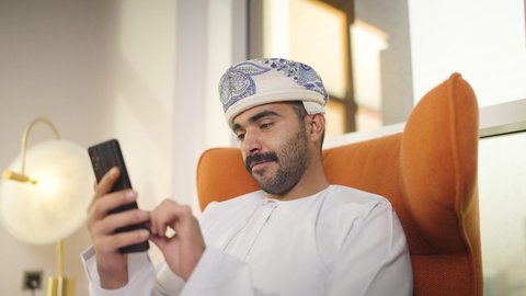 التواصل مع العملاء عن بعد، تصفح وسائل التواصل الاجتماعي ،  اجراء مكالمه فيديو ،استخدام التقنيات الحديثة و المتطورة في العمل ، مهن و أعمال في سلطنه عمان , شاب عربي خليجي عماني يرتدي الدشداشه والكمه يمسك الهاتف المحمول بيده ، يجلس على  كرسي مريح فى غرفه فندقيه , إجراء اتصال هاتفي عبر الجوال