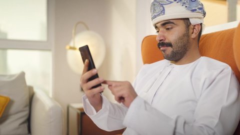 تصفح وسائل التواصل الاجتماعي ، التواصل مع العملاء عن بعد، اجراء مكالمه فيديو ،استخدام التقنيات الحديثة و المتطورة في العمل ، مهن و أعمال في سلطنه عمان , شاب عربي خليجي عماني يرتدي الدشداشه والكمه يمسك الهاتف المحمول بيده ، يجلس على  كرسي مريح فى غرفه فندقيه , إجراء اتصال هاتفي عبر الجوال