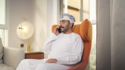 استخدام التقنيات الحديثة و المتطورة في العمل ،  تصفح وسائل التواصل الاجتماعي ، التواصل مع العملاء عن بعد، اجراء مكالمه فيديو ، مهن و أعمال في سلطنه عمان , شاب عربي خليجي عماني يرتدي الدشداشه والكمه يمسك الهاتف المحمول بيده ، يجلس على  كرسي مريح فى غرفه فندقيه , إجراء اتصال هاتفي عبر الجوال